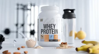 Pot de whey protéine avec un shaker et une cuillère remplie de poudre, posés sur une table avec des amandes, des flocons d'avoine et une banane, en arrière-plan léger d'équipement de gym flouté